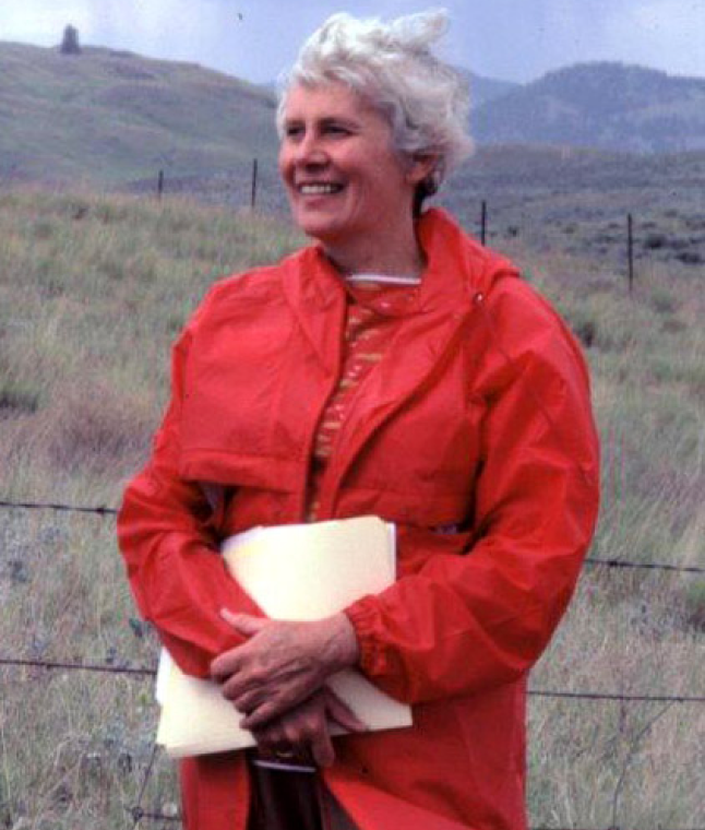 Anna Roberts, the first president of the Williams Lake Field Naturalists stands smiling in rolling grasslands with a bright red jacket.
